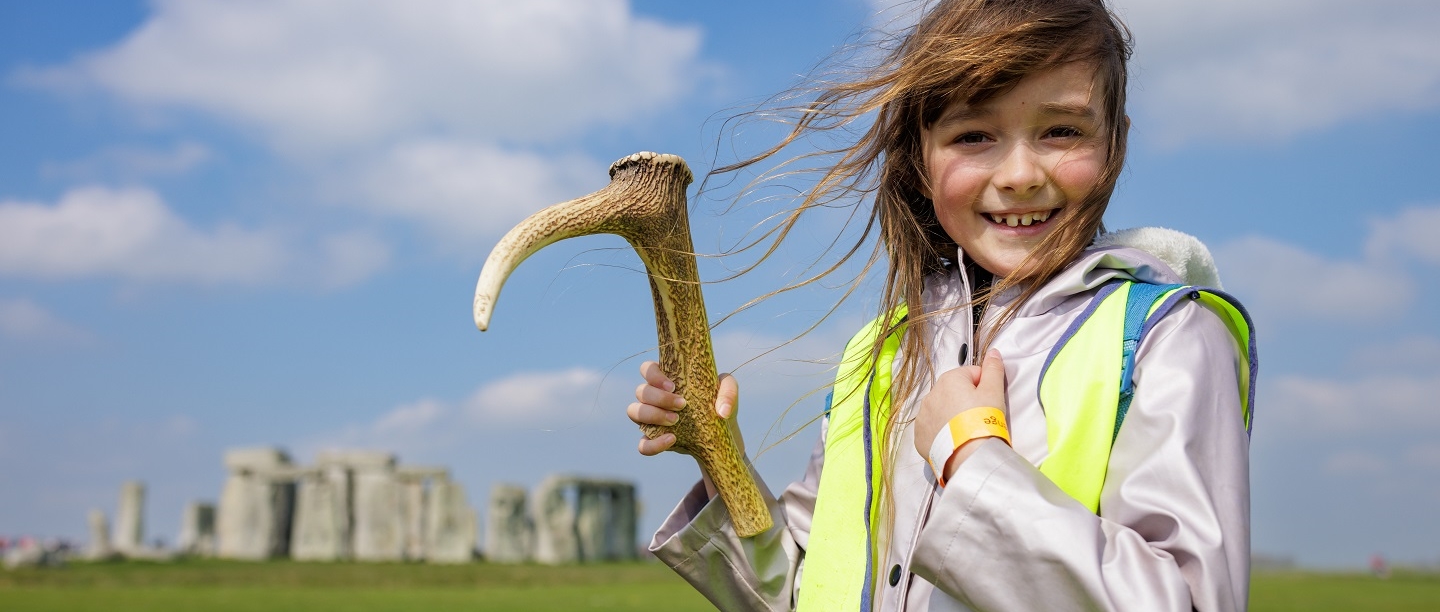  A young girl in a high vis jacket holds a replica neolithic tool in front of Stonehenge. 